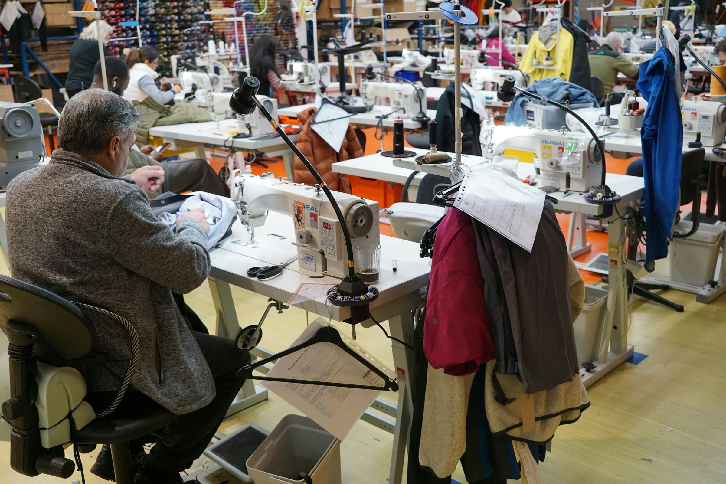 Operators at sewing machines on a busy garment floor, with jackets hanging beside them